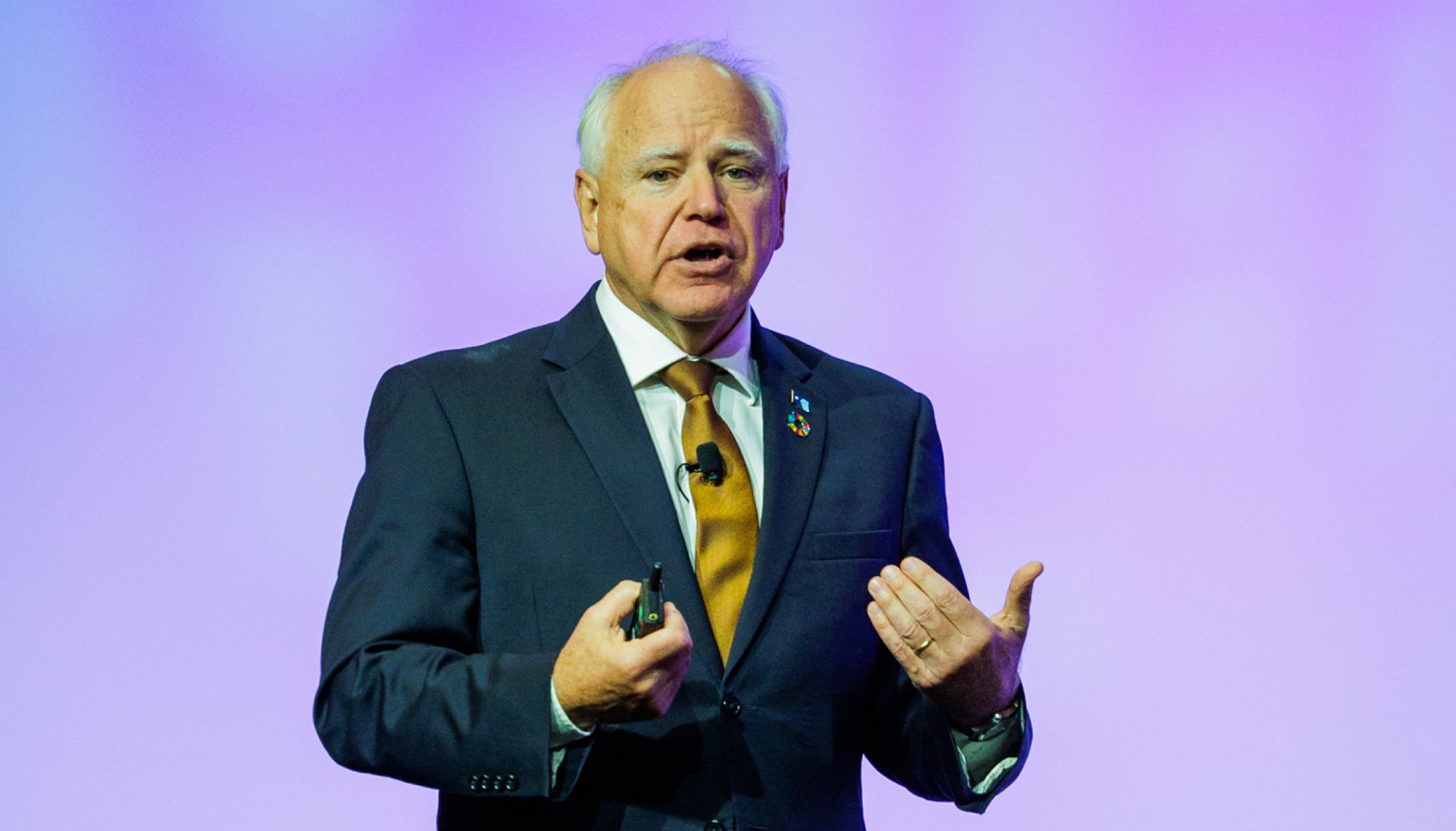 Governor Tim Walz giving a presentation wearing a dark blue suit and gold tie against a lavender backdrop