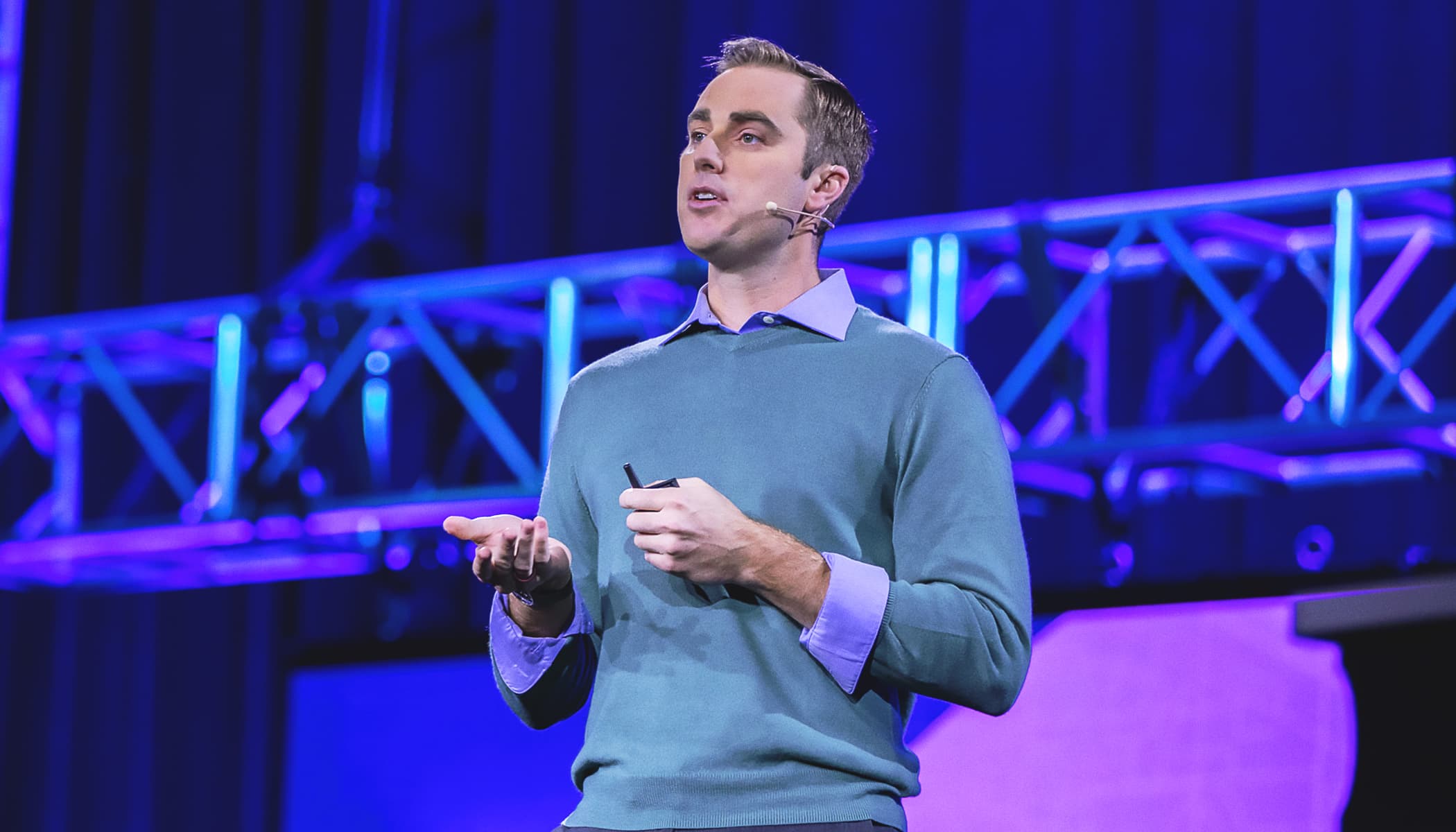 Presenter in a gray-blue sweater and lilac shirt speaks onstage against a blue curtain and metal scaffolding