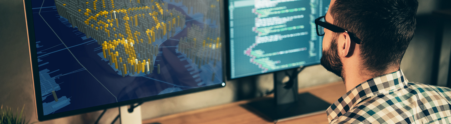 A person seated at a computer, examining a 3D visualization of map data on one monitor and software development code on another